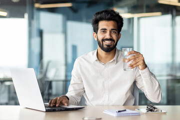An Indian young male businessman is sitting in the office at his desk and holding a glass of clean...