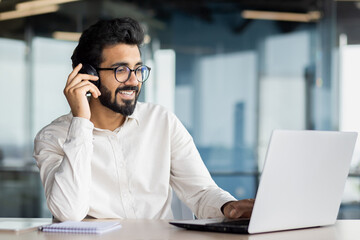 Indian young man businessman sitting in the office at the desk in front of the laptop and smilingly talking on the mobile phone