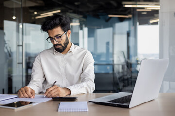 Obraz premium A focused and serious young Indian man works as an accountant, auditor, and financial analyst. Sitting at the desk in the office and working with accounts and data
