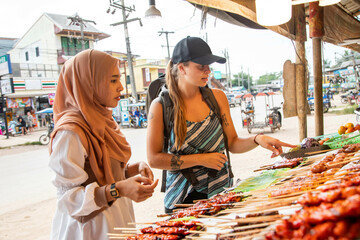 Two young women one wearing a Hijab buying street food in a rural town in Thailand
