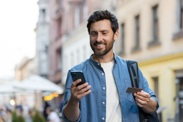 A traveling young man stands on a city street with a backpack, uses a credit card and a phone. Make...