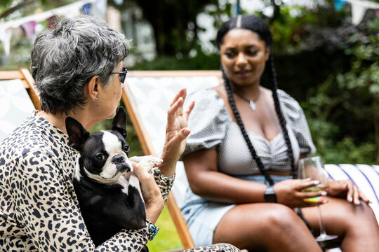 Mature white woman with a Boston terrier dog is deep in conversation with a young black woman at a garden party in the summer