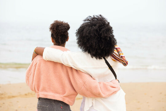 Two mixed race sisters on the beach hug each other and look out to the sea.