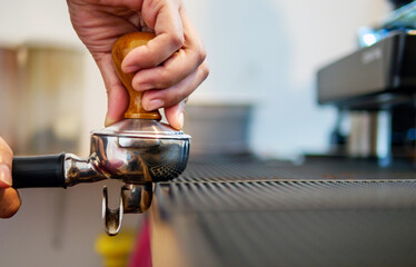 Hand of a barista holding a portafilter and a coffee tamper making an espresso coffee. Barista...