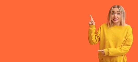 Embarrassed young woman pointing at something on orange background with space for text
