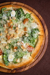 Round pizza with salad, tomatoes, cheese, croutons on wooden board. Brown background, top view.