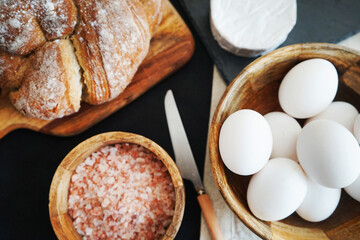 White chicken eggs in a wooden bowl next to bread, Brie cheese and coarse pink salt