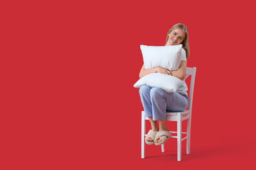 Pretty young woman in pajamas and with pillow sitting on chair against red background