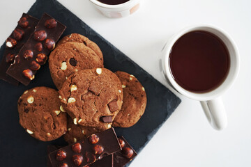 Chocolate cookies with chocolate slices and nuts on a dark stand next to two cups of tea on a white background
