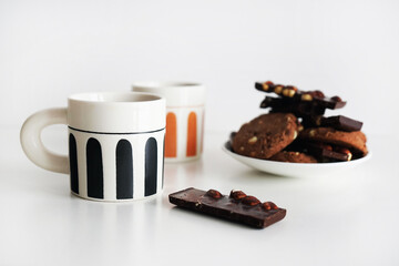 Chocolate cookies with chocolate slices and nuts on a plate next to two cups of tea on a white background