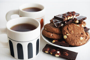 Chocolate cookies with chocolate slices and nuts on a plate next to two cups of tea on a white background
