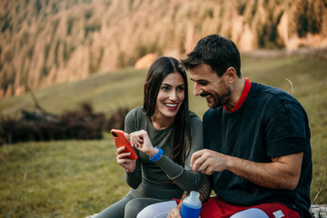 Diverse couple catching their breath after a jog on a hill road, hydrating with water and checking fitness stats on their smart devices