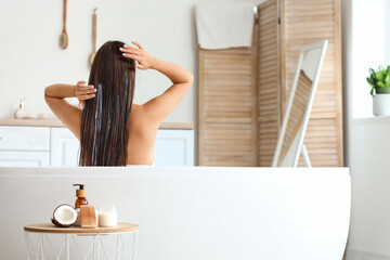 Beautiful young woman applying coconut oil on her hair in bathroom, back view