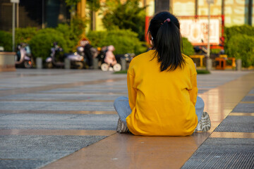 little girl in yellow sweater sitting in square with her back turned, Asian girl sitting in park...