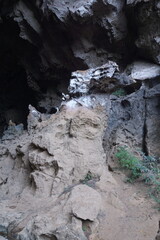 A large cave in the forest surrounded by large rocks and water flowing from the mouth of the cave.