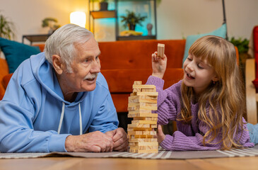 Excited little cute child girl granddaughter kid playing building block game with smiling elderly...