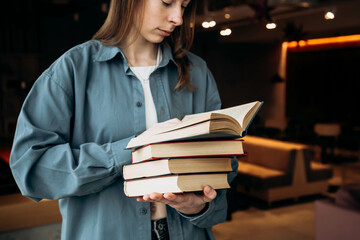 Young woman with a pile of books on a blurred library background. A stack of books in female hands. Female student in college library with books. Education, learning and reading concept.