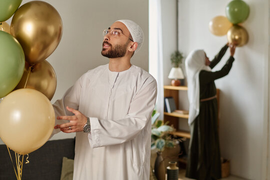 Happy young Muslim man in traditional white skullcap and kandora looking at bunch of balloons while decorating home for holiday
