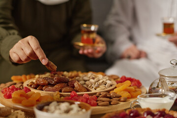 Hand of young Muslim woman taking dried date fruit from plate with variety of snacks while having festive dinner with her husband