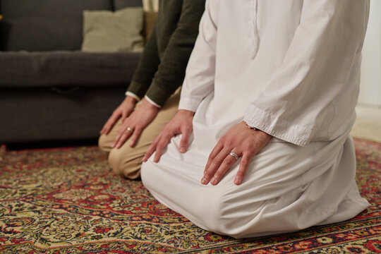 Young Muslim man in white kandora and his brother in casualwear standing on their knees on rug and performing namaz prayer