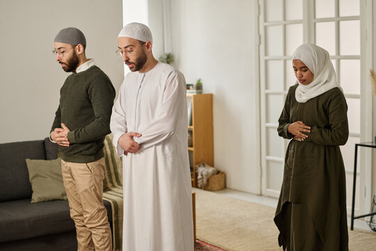 Two Muslim men and wife of one of them in traditional clothes keeping hands by abdomen while performing namaz prayer at home
