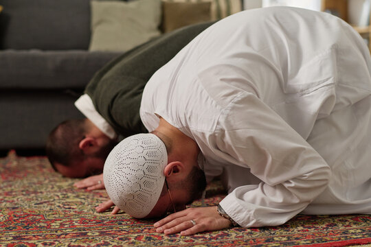 Young Muslim men in white skullcap and thobe touching carpet by head while standing on knees and bending forwards during prayer