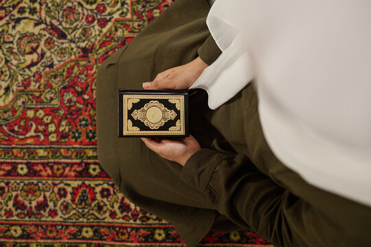 Top view of young Muslim woman in traditional dress and hijab standing on knees on the carpet and holding Holy Quran before reading it