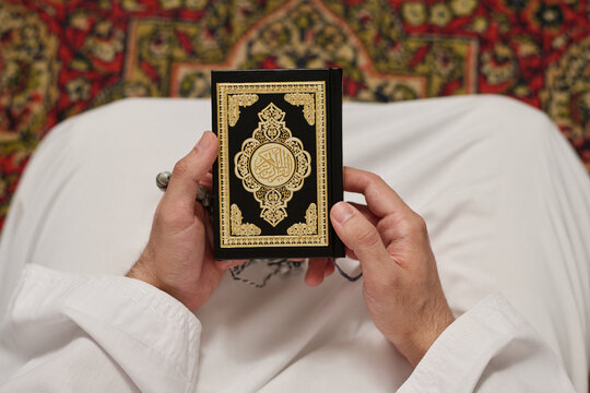Top view of Holy Quran with Arabic calligraphy and ornament on cover in hands of young unrecognizable Muslim man wearing white kandora