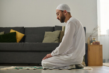 Young Muslim man in white skullcap and thobe standing on knees on rug and performing namaz prayer against couch with cushions