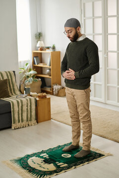 Young Muslim man in casualwear and skullcap keeping eyes closed and hands crossed by abdomen while performing namaz prayer on rug