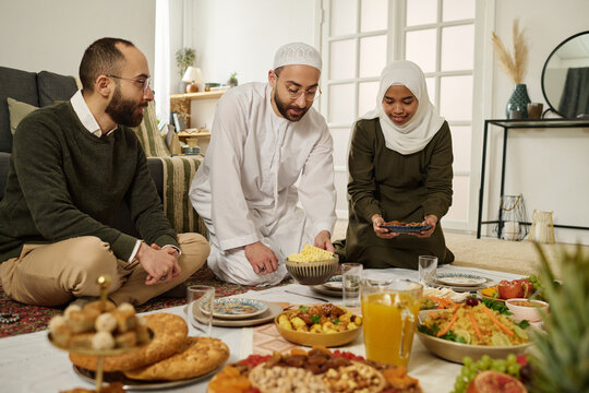 Young Muslim man in white kandora and skullcap sitting between his twin brother in casualwear and wife in hijab holding plate with snack