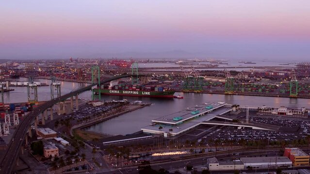 Aerial Panning Shot Of Cargo Goods In Merchant Ship Under Famous Bridge At Port Against Sky At Sunset - San Pedro, California