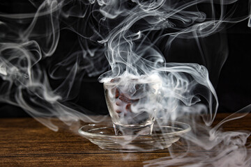 Smoke vape fog flowing out of transparent glass coffee cup filled with coffee beans on dark black background