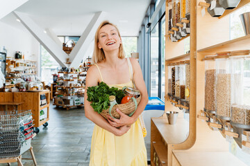 Smiling woman with basket of organic products in sustainable plastic free store. Dispensers for cereals, grains, nuts at zero waste shop.