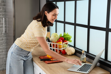 Young woman with fresh vegetables using laptop in kitchen