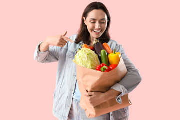 Young woman pointing at paper bag of fresh vegetables on pink background