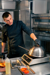 a chef in a restaurant's kitchen in a black jacket opens the lid of frying pan to check its readiness