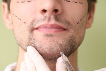 Young man with marked face receiving injection on green background, closeup. Plastic surgery concept