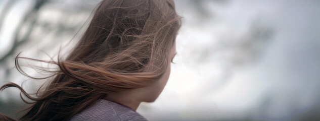 A girl looking into the blurry distance, enjoying the wind blowing her long brown hair. Rear view, outdoors, overcast. - 736107969