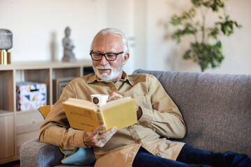 Photo of a senior man reading a book while enjoying a cup of tea, with his feet up on the sofa....
