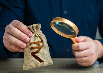A man examines a indian rupee money bag through a magnifying glass. Investigating capital origins....