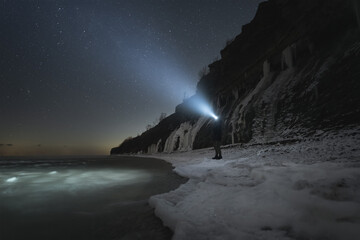 A man with a flashlight on the steep shore of the Baltic Sea in Paldiski at night in winter time.
