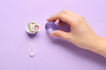 Female hand with cleaning case for contact lenses and tweezers on lilac background