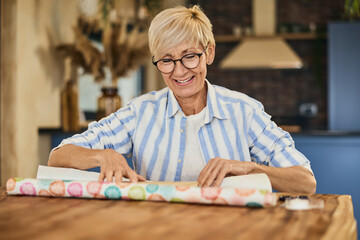 A smiling mature woman, sitting at home and packing a gift for someone.