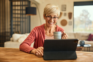 A senior woman smiling and using a digital tablet, sitting at home and holding a cup of coffee.