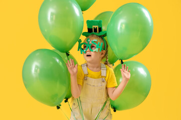 Cute little girl with leprechaun's hat, novelty glasses and balloons on yellow background. St. Patrick's Day celebration