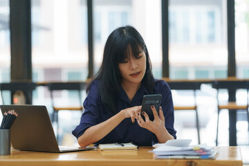 Portrait of a beautiful confident businesswoman using a laptop computer holding a mobile phone sitting in a modern office. Smiling Asian freelancer working online from home.