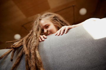 View from below. Young girl with braided hair deep leaning on sofa in well-lit living room at home....