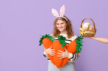 Young happy woman in bunny ears with carrot-shaped toys and hand holding Easter basket on purple background