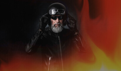 Portrait d'un motard homme avec un casque noir et une veste en cuir et des lunettes de protection et une barbe grise vintage devant un rideau de feu